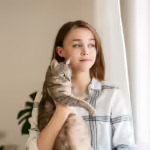 Indoor shot of amazing lady holding cat . Portrait of young woman holding cute striped cat with green eyes and looking at the window. Female hugging her cute kitty. Adorable domestic pet concept.